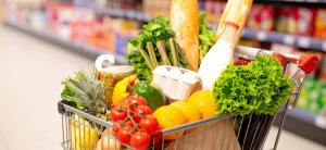 Carrinho de supermercado abastecido com frutas, verduras e alimentos frescos como alface, tomates, laranja, abacaxi, leite, ovos e pães, em corredor de mercado com prateleiras de produtos desfocadas ao fundo.